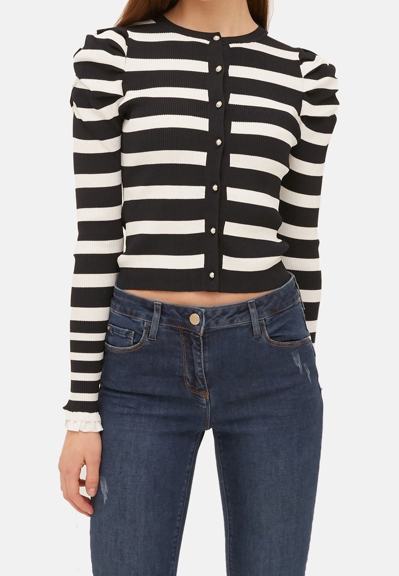 Žena oblečená v černo-bílém pruhovaném krátkém svetříku s perlovými knoflíky a vycpanými rameny, doplněná tmavě modrými džíny.