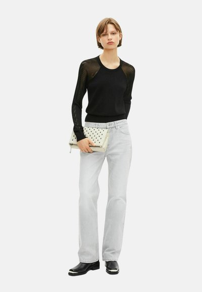 Persona joven con cabello corto, vistiendo una blusa negra de mangas de malla, jeans gris claro, zapatos negros y sosteniendo un clutch blanco con tachuelas.