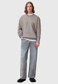 Sivý crewneck sveter s embosovaným textom, skombinovaný s voľnými svetlosivými džínsy. Model nosí čierne slip-on topánky.