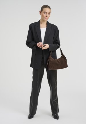 Vrouw draagt een zwarte oversized blazer en zwarte leren broek, houdt een bruine suède handtas met schouderriem vast.