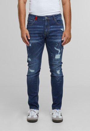 Jean en denim bleu foncé avec une coupe slim, présentant des détails déchirés, des accents bleu clair subtils et une languette rouge à la taille.