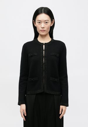 Jeune femme aux cheveux noirs et raides portant un cardigan noir boutonné avec des bordures décoratives et un pantalon noir à jambes larges, se tenant droite.