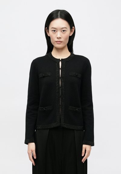 Jeune femme aux cheveux noirs et raides portant un cardigan noir boutonné avec des bordures décoratives et un pantalon noir à jambes larges, se tenant droite.