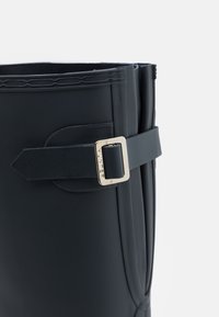 Botín de goma negro con una textura lisa, que cuenta con una correa con una hebilla plateada y un borde superior doblado como detalle de diseño.