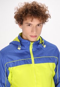 Chaqueta cortavientos azul y amarilla con cierre de cremallera, capucha ajustable y ribetes en contraste. Material ligero y resistente al agua.