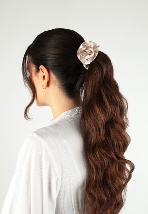 Donna con capelli castani lunghi e ondulati raccolti in una coda alta con un elastico di raso beige chiaro, che indossa una maglia bianca con texture.