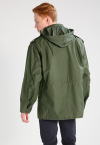 Jachetă verde măslinie cu glugă și snur, mâneci lungi și croi drept. Fabricată dintr-un material ușor, având detalii cusute.