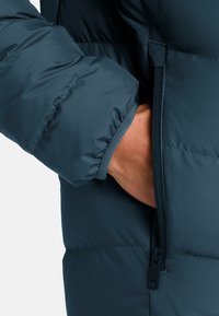Pufferjacketsleeve in donker teal, met een ritszak en elastische manchetten voor extra warmte en comfort.