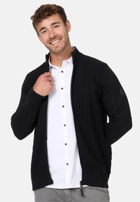 Cardigan noir zippé avec un devant en maille texturée et des manches unies. Dispose d'un col montant et d'un discret détail zippé à l'ourlet. Porté par-dessus une chemise blanche.