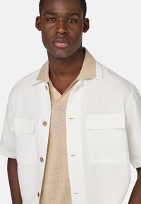 Camicia bianca a maniche corte con due taschini sul petto e colletto beige, indossata sopra una maglietta beige chiaro a scollo a V. Tessuto dalla consistenza liscia.