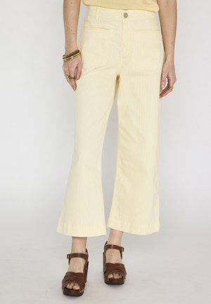 Donna che indossa pantaloni corti a righe gialle e bianche, sandali con zeppa marroni e braccialetti e anelli dorati su entrambe le mani.