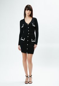 Robe noire côtelée avec un décolleté en V profond, des manches longues et des poches avant bordées de blanc. Présente des boutons blancs sur le devant.