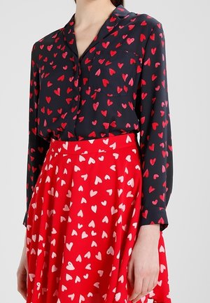 Woman wearing a black long-sleeve blouse and red skirt, both patterned with small red and white hearts.