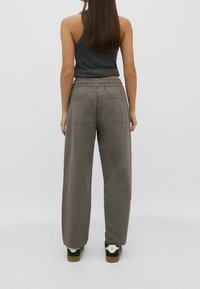 Nainen seisoo selkä kameraan päin, päällään musta hihaton paita, harmaat leveälahkeiset housut takataskuilla ja musta-valkoiset lenkkarit.