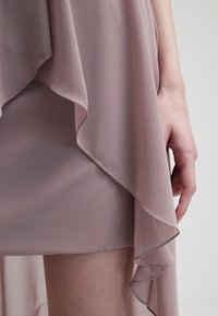 Lehounká, světle růžová šifonová látka s vrstvami volánků. Ruka drží okraj, což ukazuje na měkký, texturovaný materiál a vrstvený design.