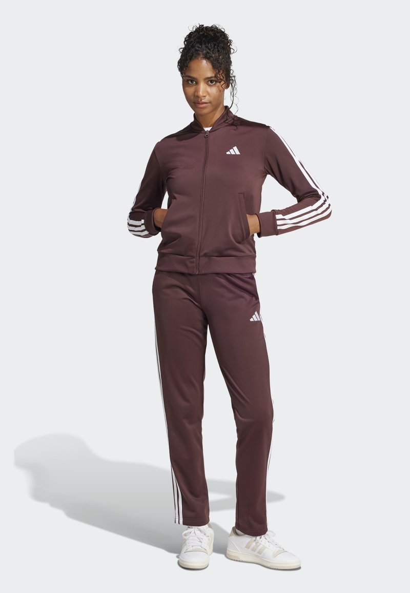 Brun jakke og bukser i sett, med hvite striper på ermene og beina, laget av glatt stoff med glidelås foran. Hvite sneakers inkludert.