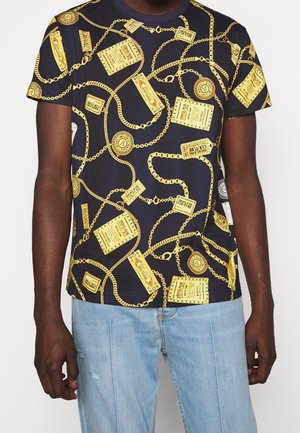 Mężczyzna w granatowej koszulce z nadrukiem złotego łańcucha i etykiety, w połączeniu z jasnoniebieskimi dżinsami na jednolitym tle.