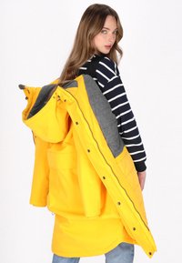 Gelbe wasserdichte Parka mit grauem Fleece-Futter, Druckknöpfen und Kapuze, getragen über einem schwarz-weißen gestreiften Shirt.
