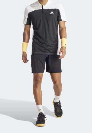 Camiseta deportiva en blanco y negro de manga corta, combinada con pantalones cortos negros. Lleva puestas muñequeras amarillas y zapatos negros con suelas amarillas.