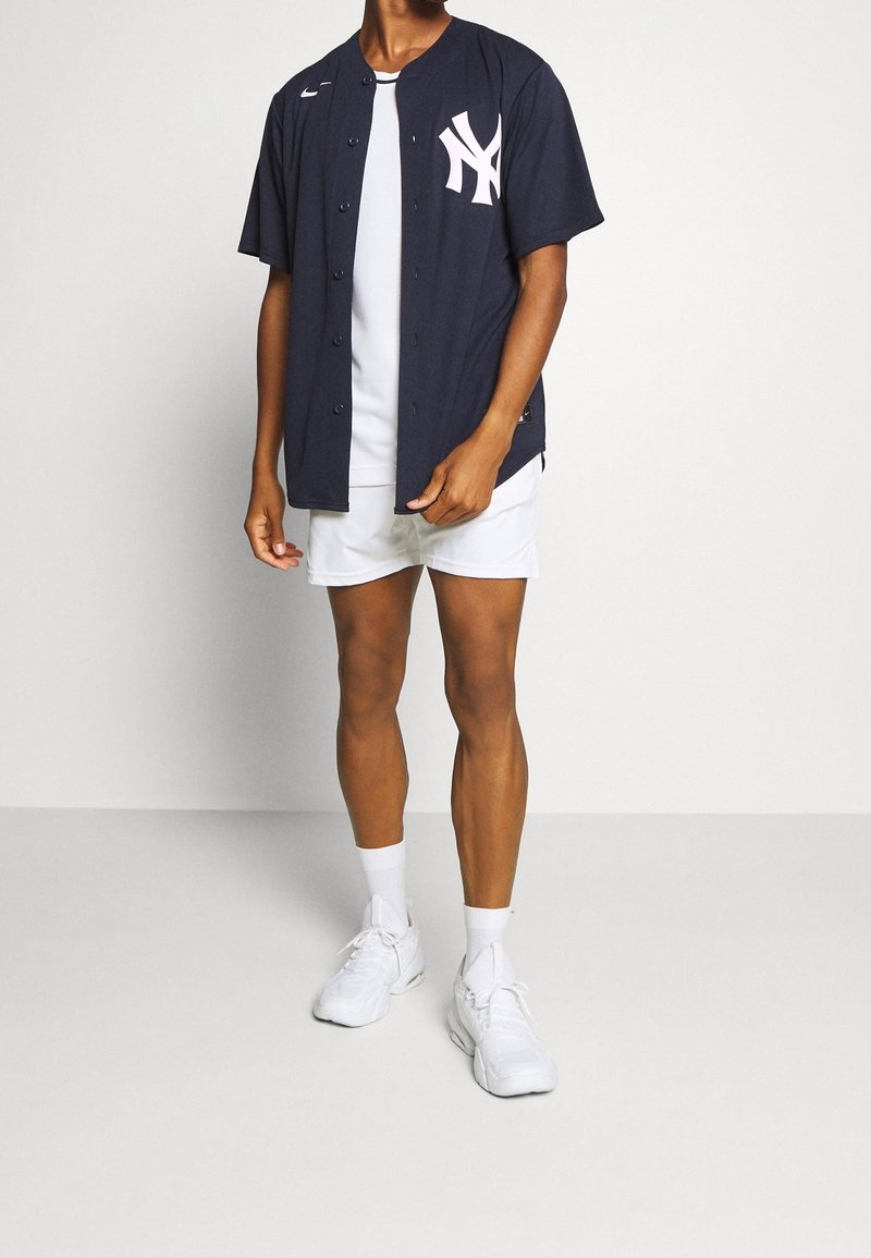 Tummansininen baseball-paita, jossa on lyhyet hihat, napit edessä ja vaaleanpunainen NY-logo. Päällä valkoisen paidan kanssa, yhdistetty valkoisiin shortseihin ja kenkiin.