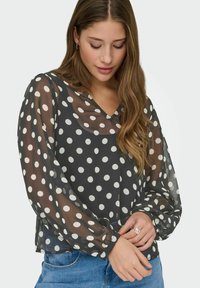 Femme aux longs cheveux bruns portant une blouse noire transparente à pois blancs et un jean bleu, regardant vers le bas avec les mains jointes.