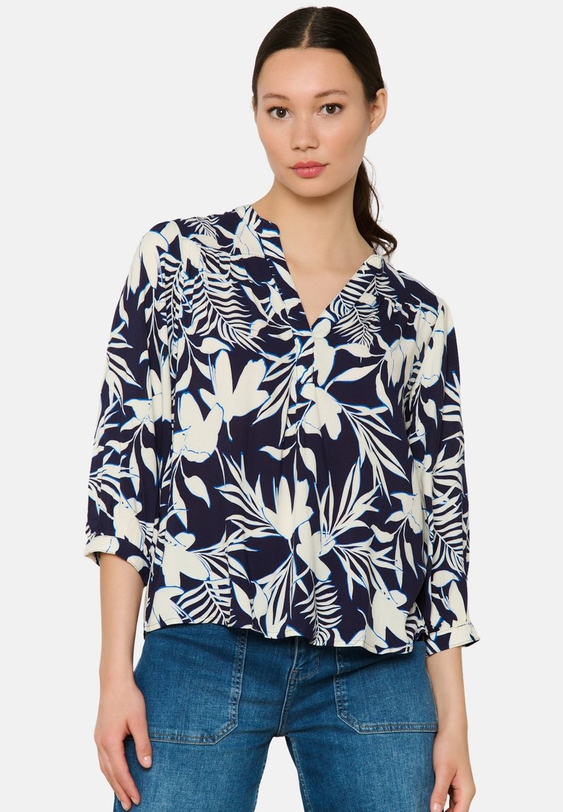 Marineblaue Bluse mit Blumenmuster, Dreiviertelärmeln, V-Ausschnitt und leichtem Stoff. Kombiniert mit blauen Jeanshosen.