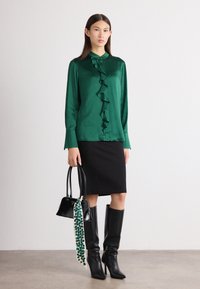 Grüne Satinbluse mit gerafftem Vorderteil, schwarzer knielanger Rock, schwarze kniehohe Stiefel und eine schwarze Handtasche mit einem gemusterten Schal-Akzent.