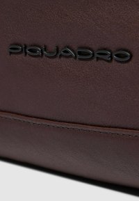Prodotto in pelle marrone con una texture liscia, cuciture evidenziate e un prominente logo nero "Piquadro".