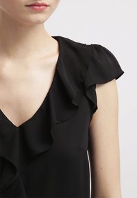 Zbliżenie na osobę w czarnej bluzce bez rękawów z falbaniastymi detalami wzdłuż dekoltu i ramienia.
