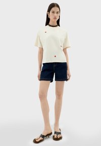 Witte t-shirt met korte mouwen en rode geborduurde bloemen, gecombineerd met donkere denim shorts en zwarte sandalen met bruine bontaccenten.