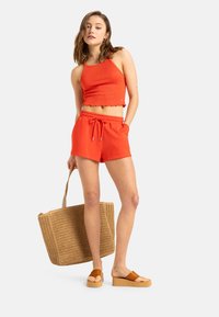 Helles rotes, kurzes, geripptes Tanktop und passende Shorts mit Kordelzug, kombiniert mit einer gewebten beigefarbenen Tragetasche und beigen Plattform-Sandalen.