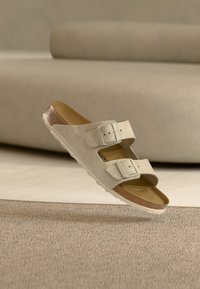 Μπεζ σουέτ σανδάλι με δύο ρυθμιζόμενες αγκράφες και πάτο από φελλό, που αιωρείται πάνω από μπεζ υφασμάτινο χαλί σε έναν χώρο με ουδέτερους τόνους.