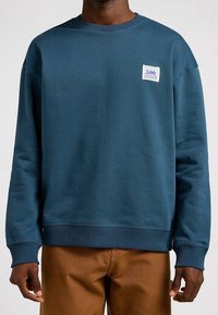 Sudadera de algodón de color azul marino con puños y dobladillo acanalados, que presenta un parche de logo blanco en el pecho y un diseño de cuello redondo.