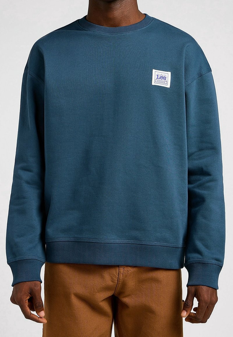 Sudadera de algodón de color azul marino con puños y dobladillo acanalados, que presenta un parche de logo blanco en el pecho y un diseño de cuello redondo.