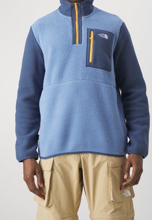 Homme portant un pull polaire bleu clair et bleu foncé avec une fermeture éclair jaune, et un short cargo beige avec des poches.