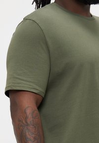 Tricou verde măslinie cu mâneci scurte, realizat dintr-un material moale, cu gât rotund. Detalii de cusătură și textură netedă; purtat cu un tatuaj pe braț vizibil.