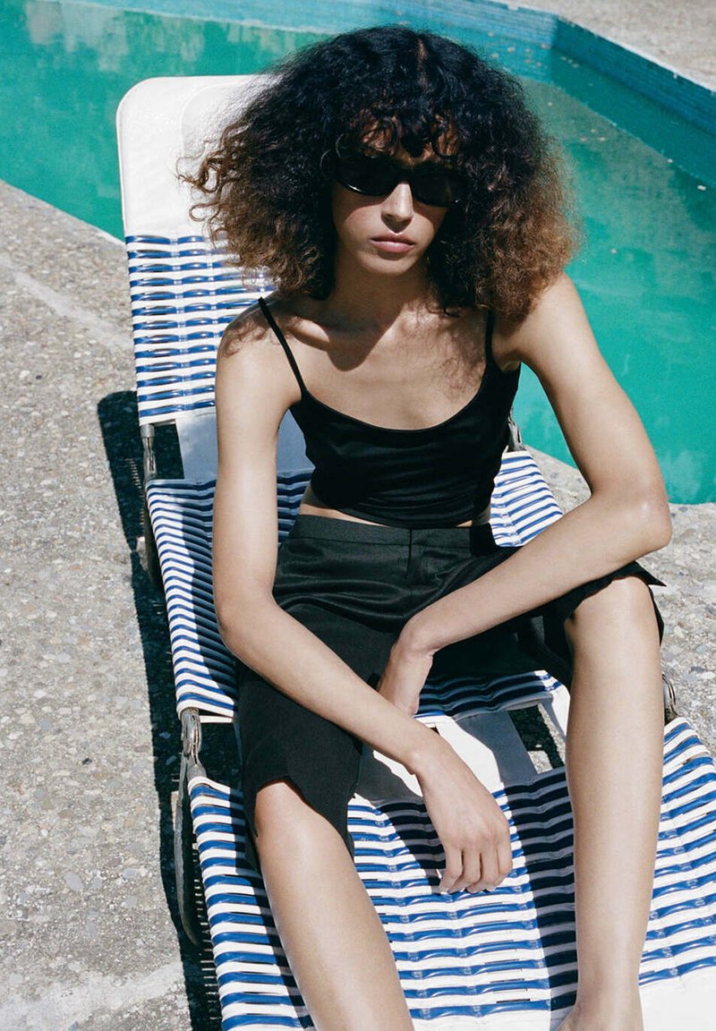 Jeune personne aux cheveux bouclés et lunettes de soleil assise sur une chaise longue à rayures bleues près d'une piscine, vêtue de noir.