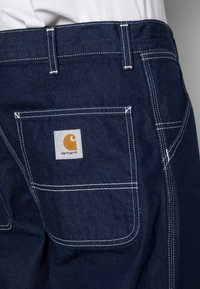 Pantalones de trabajo de mezclilla oscura con costuras blancas, con un bolsillo trasero y un pequeño parche con el logo de Carhartt en el bolsillo derecho.