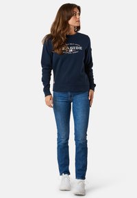 Navy sweatshirt met een ronde hals, met witte grafische tekst en een klein logo. Gecombineerd met blauwe jeans en witte sneakers.