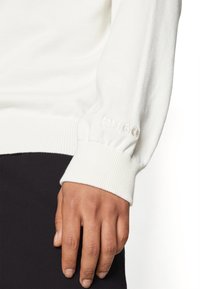 Gros plan d'une main portant un pull blanc à manches longues avec un poignet côtelé brodé de "HUGO" près du poignet et un pantalon noir.