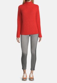 Maglione a collo alto rosso a maglia con maniche lunghe, abbinato a jeans grigi chiari con una striscia laterale e sneakers con dettagli arancioni.