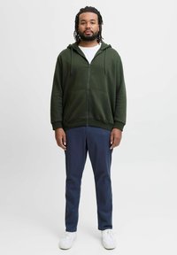 Groene rits-hoodie met trekkoorden, voorzakken, en een wit T-shirt eronder, gecombineerd met marineblauwe broek en witte sneakers.