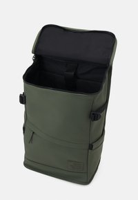 Sac à dos étanche vert avec un dessus zippé, un intérieur rembourré, une poche extérieure et des bretelles réglables ; possède une surface texturée et un patch avec logo.