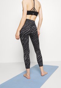 Frau, die barfuß auf einer blauen Yogamatte steht, trägt einen schwarzen Sport-BH und schwarze-graue Leggings mit Zebra-Muster, mit dem Rücken zur Kamera gewandt.