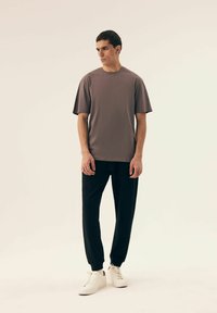 Szara koszulka z krótkim rękawem o klasycznym dekolcie, w połączeniu z czarnymi zwężającymi się dresami i białymi sneakersami, na neutralnym tle.