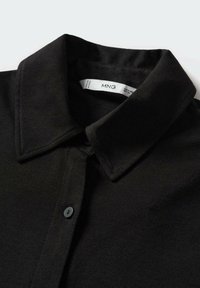 Chemise noire à col avec une patte à boutons, montrant le bouton supérieur fermé et une étiquette blanche visible à l’intérieur de l’encolure avec le texte de la marque.