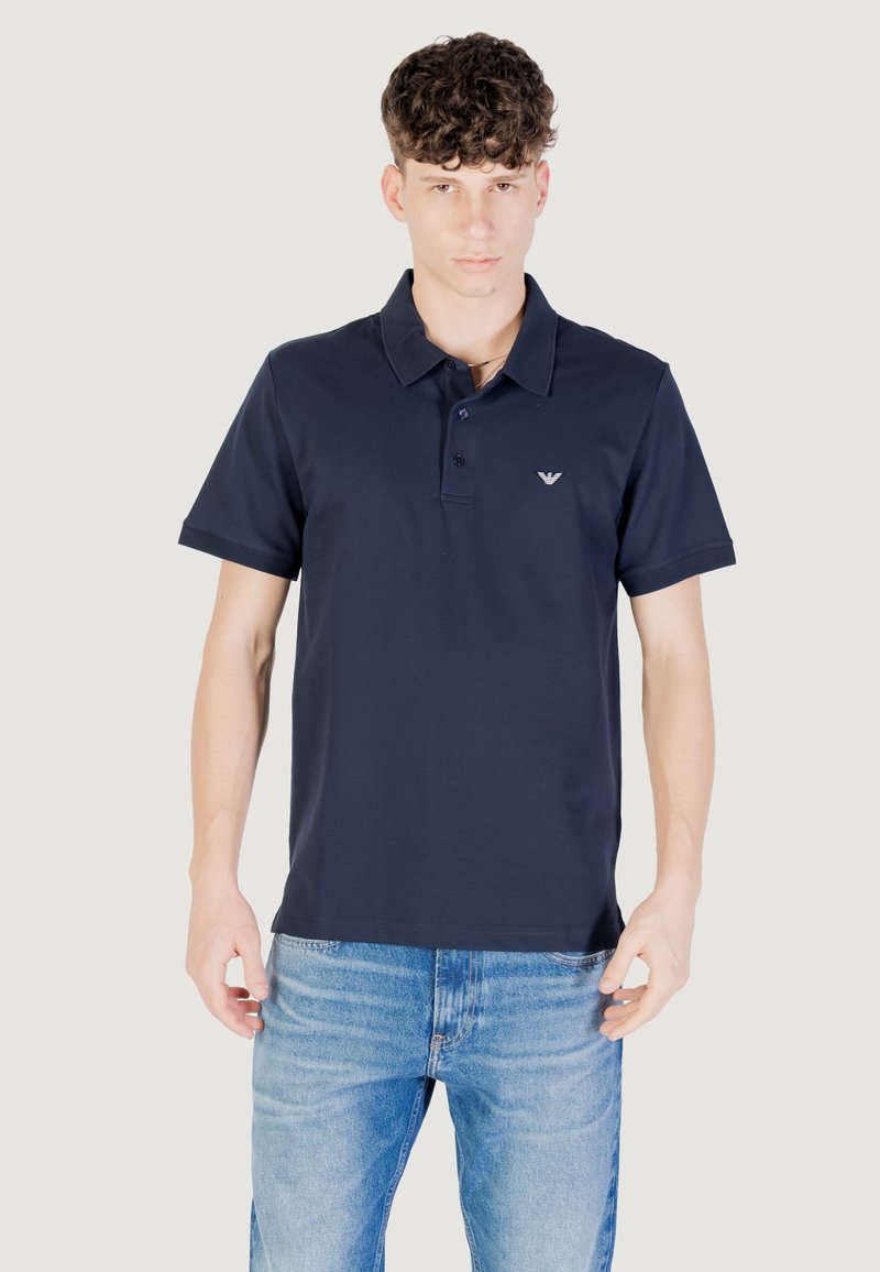 Temno moder polo majica s ovratnikom, kratkimi rokavi in majhnim logotipom na prsih. Narejena iz gladkega, lahkega materiala.