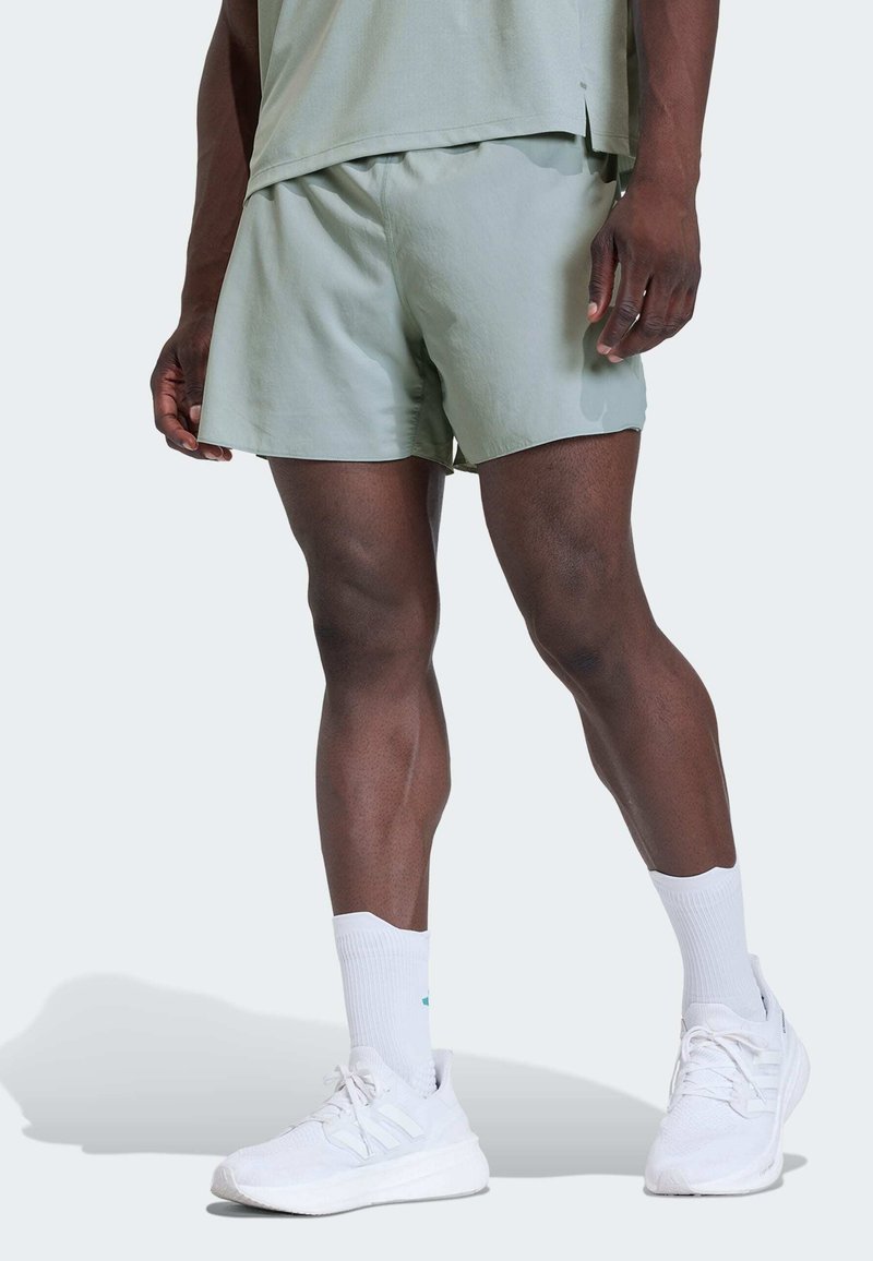 Άνδρας που φοράει ανοιχτό πράσινο σορτς, λευκές κάλτσες και λευκά αθλητικά παπούτσια, στέκεται σε φωτεινό φόντο.