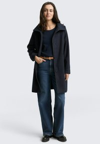Marineblauwe wollen blend jas met grote kraag, knopen aan de voorkant en zijzakken, gecombineerd met donkerblauwe flared jeans en zwarte schoenen.