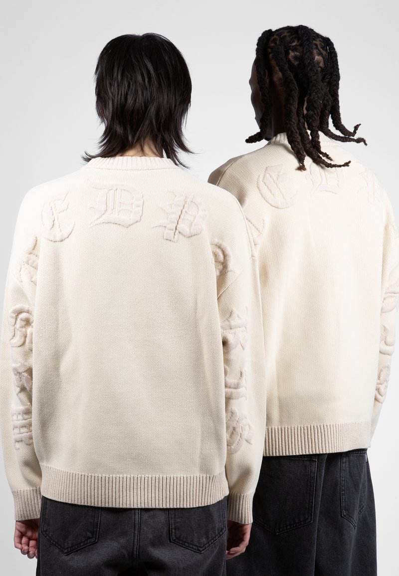 Deux personnes vues de dos portant des pulls en maille crème avec des lettres gothiques en relief, associés à un pantalon noir, sur un fond uni.